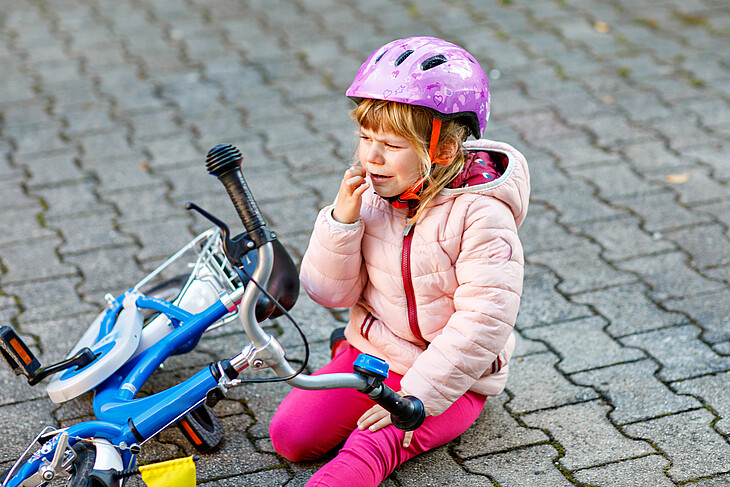 Cute little girl sitting on the ground after falling off her bike. Upset crying preschool child with safe helmet getting hurt while riding a bicycle. Active family leisure with kids.